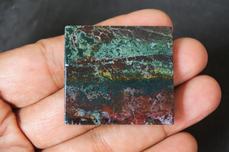 May include: A green, brown and red banded agate cabochon with a polished surface. The stone has a layered appearance with swirls of colour.