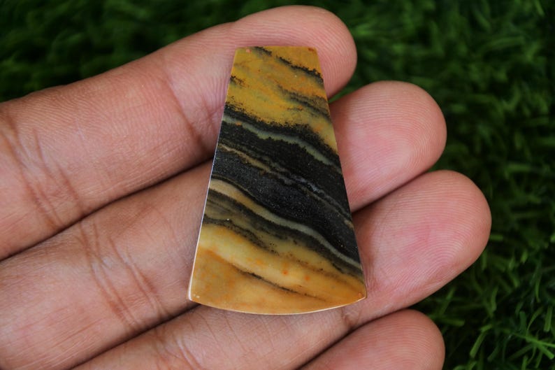 May include: A yellow and black striped cabochon gemstone with a smooth, polished surface. The gemstone is shaped like a triangle with rounded edges.