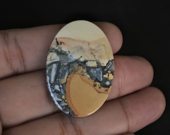 May include: Oval shaped, polished, natural stone cabochon with a beige, brown, gray, and black pattern.