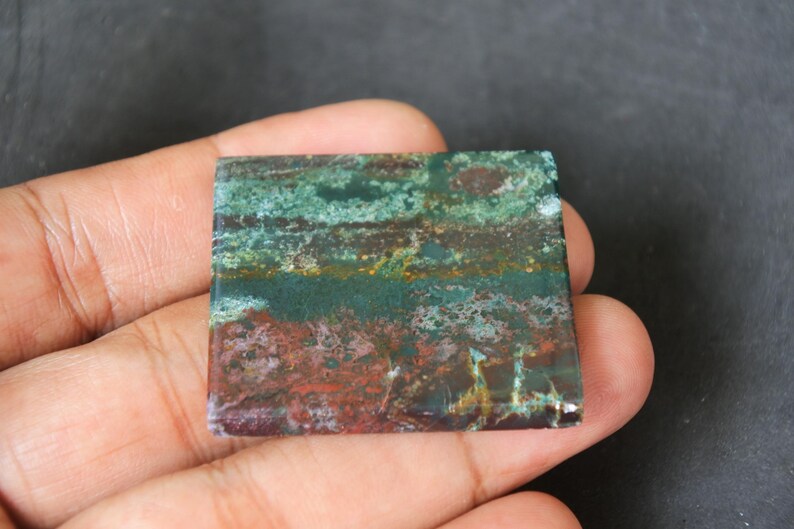 May include: A square, green, brown, and white patterned stone cabochon. The stone has a layered, banded appearance with a variety of colours and textures.