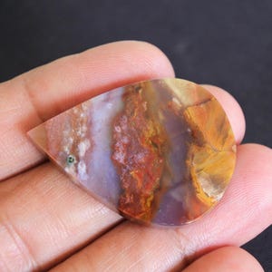 May include: A polished, teardrop-shaped gemstone displaying a mix of colours. The stone has red, orange, yellow, and purple hues, with a small green inclusion. The gemstone is held in a hand.