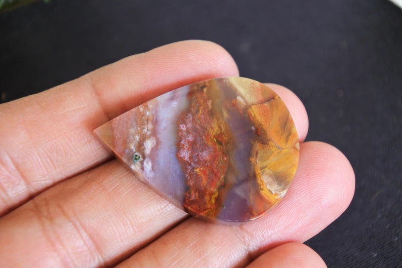 May include: A polished, teardrop-shaped gemstone displaying a mix of colours. The stone has red, orange, yellow, and purple hues, with a small green inclusion. The gemstone is held in a hand.
