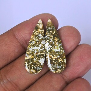 May include: A pair of teardrop-shaped green and white agate cabochons with a speckled pattern. The cabochons are held in a hand.