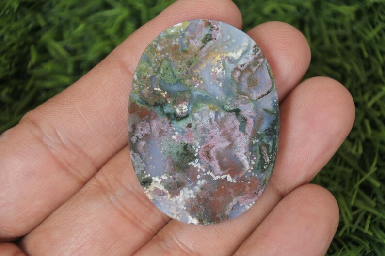 May include: A close-up of a colorful agate cabochon with a mix of green, pink, white, and brown colors. The cabochon is oval shaped and has a smooth, polished surface.