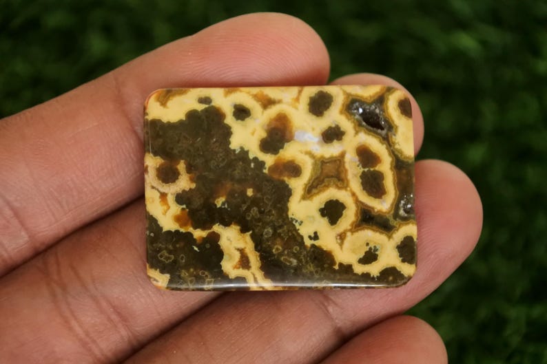 May include: A rectangular gemstone with a unique pattern of dark brown spots and swirls on a light yellow background. The polished stone is held in a hand, showcasing its natural beauty and intricate design. This stone is ideal for jewelry making.