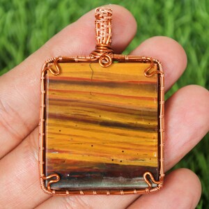 Op de afbeelding: Vierkante hanger met een koperdraadframe en balein. De hanger heeft een rechthoekige steen met horizontale banden van geel, bruin en oranje. De steen is een natuurlijk materiaal, mogelijk jaspis of agaat.