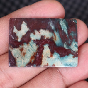 May include: A rectangular, polished stone with a swirling pattern of red, white, and blue colors. The stone has a smooth, polished surface and is suitable for jewelry making or crafting.