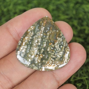 Puede incluir: Una piedra preciosa pulida con forma de lágrima. La piedra presenta una mezcla de colores verdes, marrones y blancos, con un patrón moteado. La piedra preciosa se sostiene en una mano, con un fondo verde borroso.