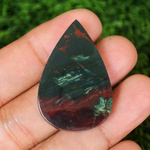 May include: A teardrop-shaped cabochon of green, red, and brown moss agate. The stone has a polished finish and is set in a silver bezel.