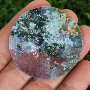 May include: A circular, polished stone displaying a natural pattern of colours. The stone has a mix of green, blue, red, and white hues, creating an artistic design. The stone is held in a hand, showing its size and texture.