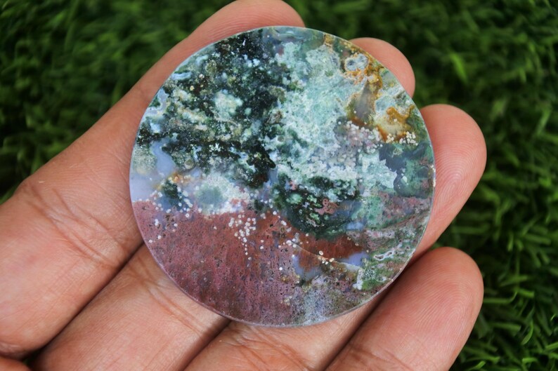 May include: A circular, polished stone displaying a natural pattern of colours. The stone has a mix of green, blue, red, and white hues, creating an artistic design. The stone is held in a hand, showing its size and texture.