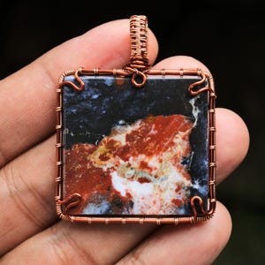 Puede incluir: Un colgante envuelto en alambre de cobre con una piedra de ágata cuadrada multicolor con patrones rojos, blancos y negros.