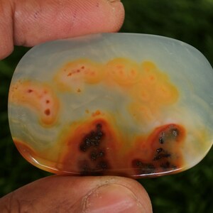 Pode incluir: Uma pedra preciosa polida em forma oval, com uma base azul p&aacute;lido transl&uacute;cida. A pedra apresenta padr&otilde;es em espiral de laranja, vermelho e castanho escuro, criando um design natural &uacute;nico. A pedra preciosa &eacute; segurada por uma m&atilde;o.