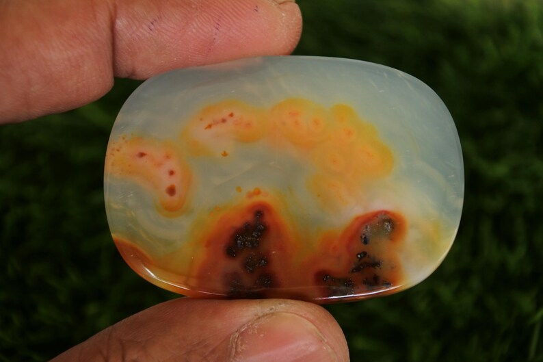 Pode incluir: Uma pedra preciosa polida em forma oval, com uma base azul p&aacute;lido transl&uacute;cida. A pedra apresenta padr&otilde;es em espiral de laranja, vermelho e castanho escuro, criando um design natural &uacute;nico. A pedra preciosa &eacute; segurada por uma m&atilde;o.