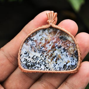 Puede incluir: Un colgante envuelto en alambre de cobre que presenta una piedra natural única con patrones marrones, blancos y negros.