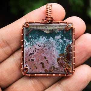 Może przedstawiać: Zawieszka owinięta miedzianym drutem z kwadratowym, wielokolorowym agatem w odcieniach różowego, zielonego i brązowego.