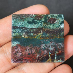 May include: A square piece of green, brown, and red jasper with a layered pattern. The stone has a polished surface and is held in a hand.