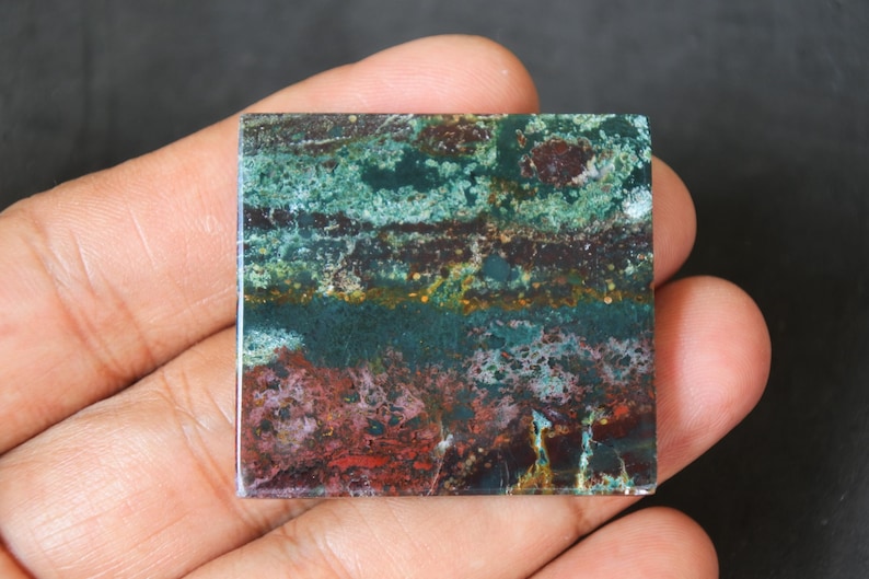 May include: A square piece of green, brown, and red jasper with a layered pattern. The stone has a polished surface and is held in a hand.