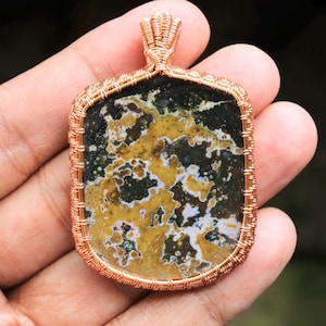 Puede incluir: Un colgante envuelto en alambre de cobre con una piedra de ágata de forma cuadrada, multicolor, con remolinos marrones, amarillos, blancos y negros.