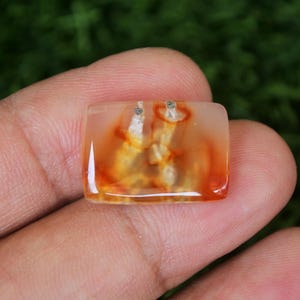 Peut inclure: Une pierre précieuse rectangulaire et translucide avec une palette de couleurs chaudes d'orange, de jaune et de blanc. Deux petits clous en argent sont fixés sur le dessus de la pierre. La pierre est tenue dans une main sur un fond vert flou.