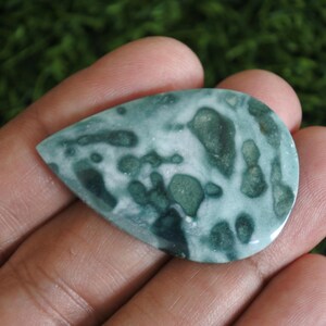 May include: A teardrop-shaped polished gemstone featuring a mottled pattern of deep forest green and creamy white patches. The stone has a smooth, reflective surface with organic, irregular shapes distributed across its entire face.