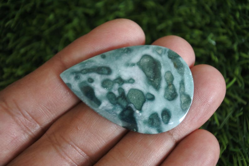 May include: A teardrop-shaped polished gemstone featuring a mottled pattern of deep forest green and creamy white patches. The stone has a smooth, reflective surface with organic, irregular shapes distributed across its entire face.