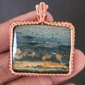 Pode incluir: Um pingente envolto em fio de cobre com uma pedra de jaspe quadrada verde, marrom e dourada.