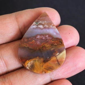 May include: A teardrop-shaped gemstone with a landscape pattern. The stone displays layers of brown, orange, purple, and white. The polished surface reflects light, highlighting the natural variations in the stone. A unique piece for jewellery.