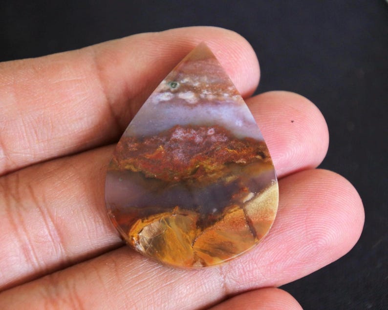 May include: A teardrop-shaped gemstone with a landscape pattern. The stone displays layers of brown, orange, purple, and white. The polished surface reflects light, highlighting the natural variations in the stone. A unique piece for jewellery.