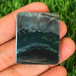 May include: A green, gray, and white agate cabochon with a layered pattern. The cabochon is rectangular and has a smooth, polished surface.