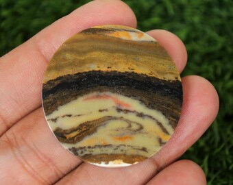 May include: A circular gemstone with a layered pattern of colours. The stone displays bands of yellow, black, brown, and cream, with hints of orange and pink. The stone is held in a hand against a green background.