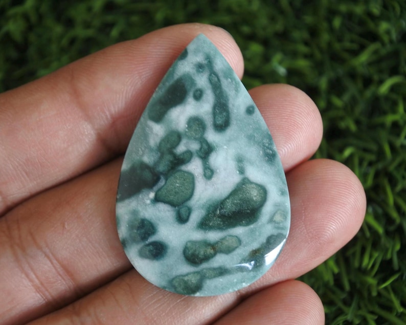 May include: A teardrop-shaped polished gemstone featuring a mottled pattern of deep forest green and creamy white patches. The stone has a smooth, reflective surface with irregular organic shapes distributed across its entire face.