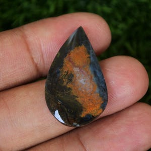 Pode incluir: Uma pedra preciosa em cabochão em forma de lágrima com um padrão azul, marrom e laranja. A pedra tem uma superfície lisa e polida.