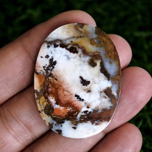 May include: An oval-shaped polished gemstone featuring a marbled pattern of white, brown, tan, and orange colors. The stone displays intricate dark brown dendritic inclusions and translucent patches throughout its smooth, glossy surface.