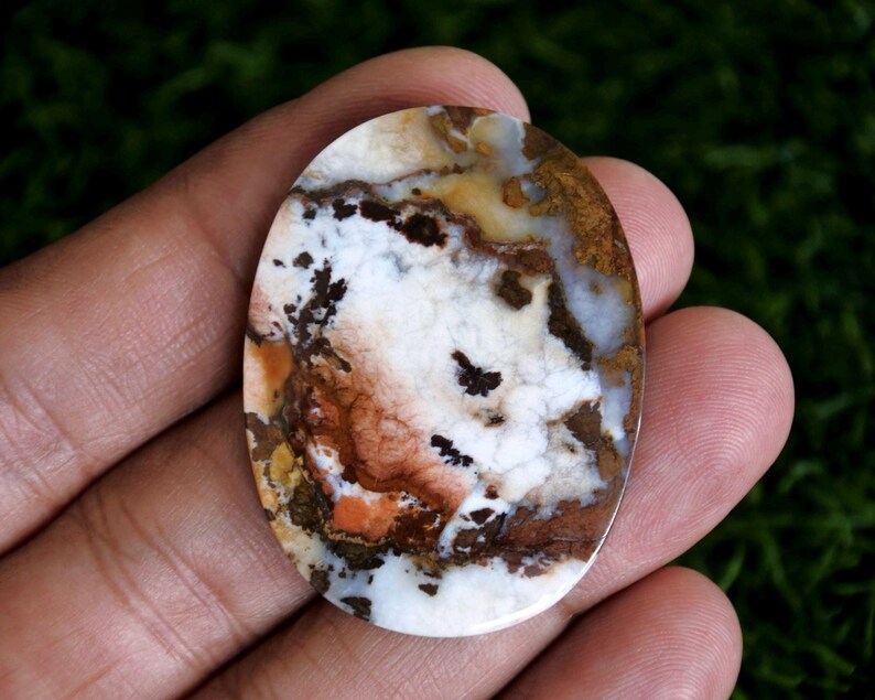 May include: An oval-shaped polished gemstone featuring a marbled pattern of white, brown, tan, and orange colors. The stone displays intricate dark brown dendritic inclusions and translucent patches throughout its smooth, glossy surface.