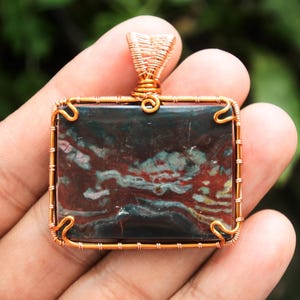 Puede incluir: Un colgante envuelto en alambre de cobre con una piedra de ágata cuadrada multicolor con remolinos rojos, blancos y negros.
