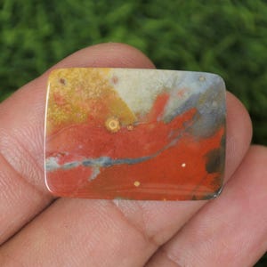May include: A red, yellow and white agate cabochon with a mottled pattern. The stone has a smooth, polished surface.