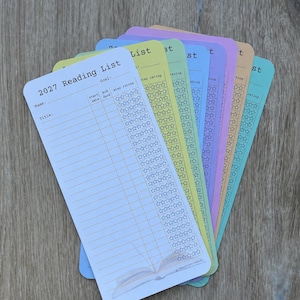 May include: A stack of rectangular 2027 Reading List bookmarks in pastel colours including white, yellow, blue, purple, orange, and teal. Each bookmark features fields for name, title, goal, start date, end date, and a column of star rating icons.