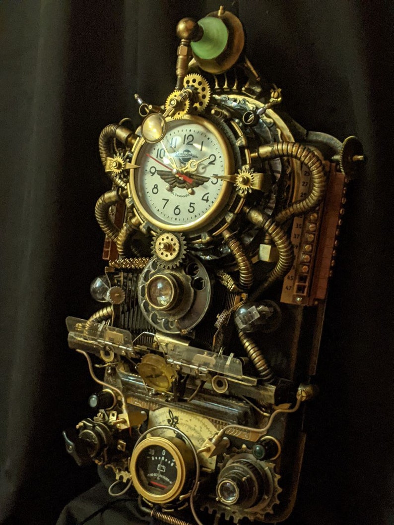 Puede incluir: Un reloj de pared de estilo steampunk con una esfera crema, n&uacute;meros negros y un emblema de ave estilizado. El reloj est&aacute; encerrado en una compleja disposici&oacute;n de engranajes, tubos y medidores de lat&oacute;n, con un elemento de vidrio verde en la parte superior. El dise&ntilde;o es intrincado e industrial.
