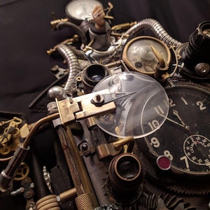 May include: A steampunk-style clock with multiple gears, dials, and lenses. The clock face has Roman numerals and a red accent. The clock is made of metal and has a brass finish.