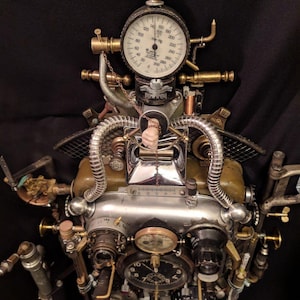May include: A steampunk-style sculpture made of metal and other materials. It features a variety of gauges, dials, and pipes, and has a hand-like object in the center. The sculpture is painted in shades of silver, gold, and brown.