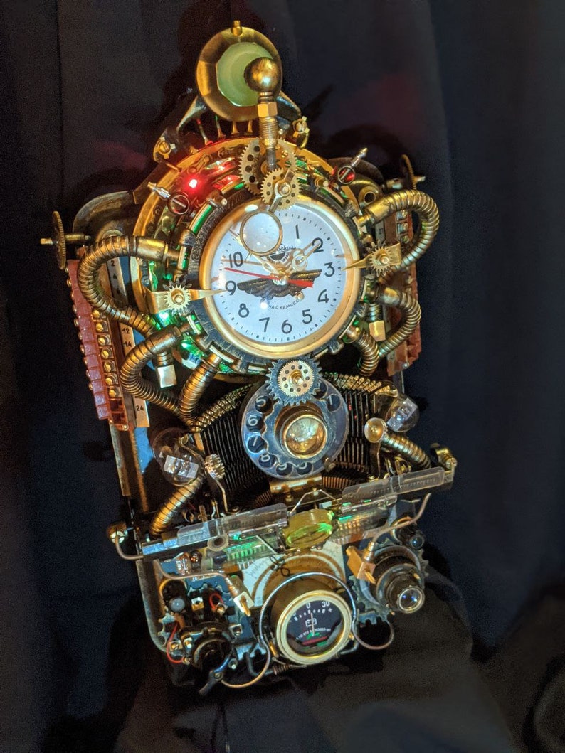 Puede incluir: Un reloj de estilo steampunk con una esfera blanca, engranajes dorados y tuber&iacute;as de cobre. El reloj tiene una est&eacute;tica vintage con detalles intrincados, incluyendo un medidor y elementos decorativos. El reloj est&aacute; iluminado con luces verdes y rojas.