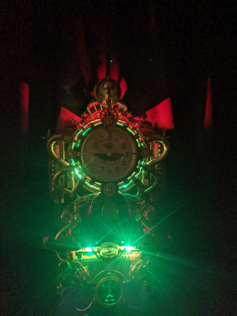 Puede incluir: Reloj de estilo steampunk con esfera redonda y detalles intrincados en lat&oacute;n y cobre. El reloj est&aacute; iluminado con luces LED verdes y rojas, creando un efecto dram&aacute;tico. El dise&ntilde;o incorpora engranajes, tubos y otros elementos mec&aacute;nicos.