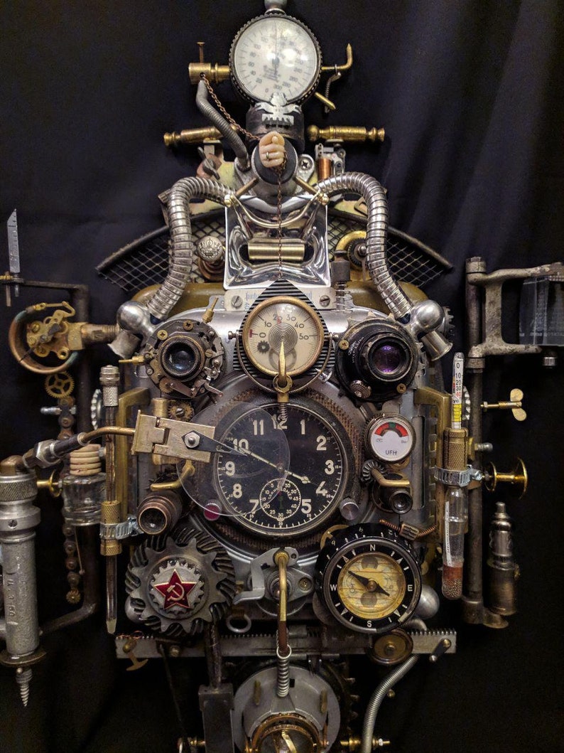 May include: A steampunk-style clock with a variety of gears, dials, and gauges. The clock face is surrounded by a metal frame with a red star and a compass. The clock is made of metal and has a distressed finish.