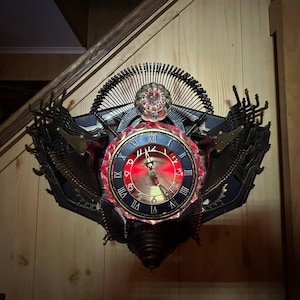 May include: A unique wall clock with a steampunk aesthetic. The clock features a round face with Roman numerals, a red illuminated center, and is surrounded by typewriter keys and metal embellishments. The clock is mounted on a wooden wall.
