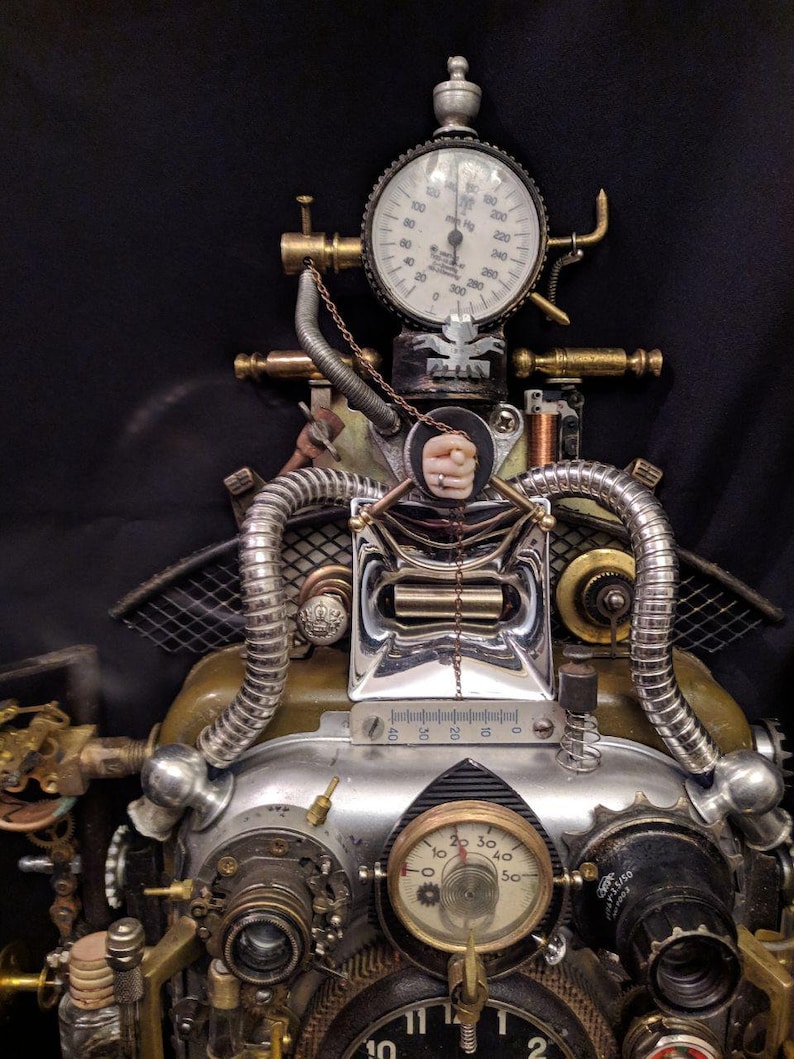 May include: A steampunk-style clock with a silver and brass finish. The clock has a variety of gears, pipes, and gauges, including a pressure gauge with the text "000200" and a smaller gauge with the text "20 30 40 50".