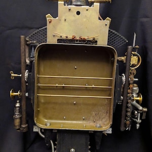 May include: A steampunk-style robot sculpture made of metal and brass. The robot has a rectangular body with a grid pattern and a round head with a gear-like design. The robot is standing on a black surface.