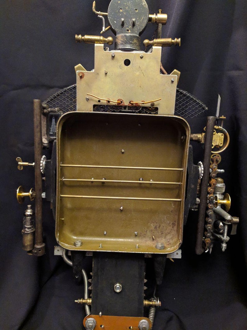 May include: A steampunk-style robot sculpture made of metal and brass. The robot has a rectangular body with a grid pattern and a round head with a gear-like design. The robot is standing on a black surface.