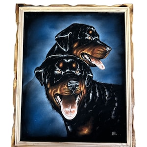 May include: A framed black velvet painting of two Rottweiler dogs. The dogs are facing forward with their tongues out. The background is a dark blue colour.