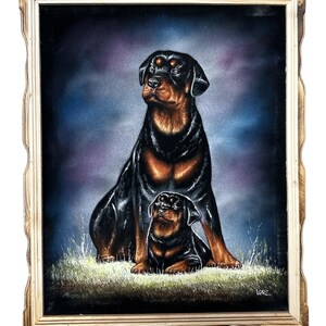 May include: A framed black velvet painting of two Rottweilers, one adult and one puppy, sitting in a field. The adult dog is looking up and the puppy is looking at the viewer. The background is a dark blue and purple gradient.